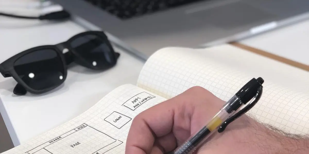 A hand sketches a website layout and notes in a grid notebook beside a laptop and sunglasses on a desk.