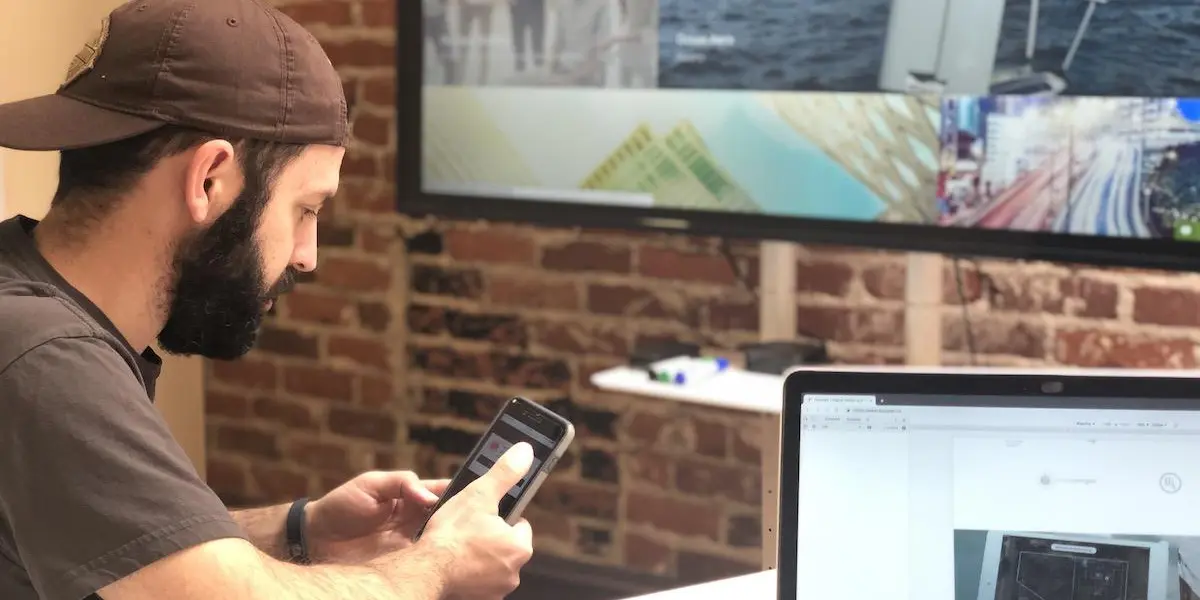 A person interacts with a mobile app while seated at a desk with a laptop and a presentation screen in the background.